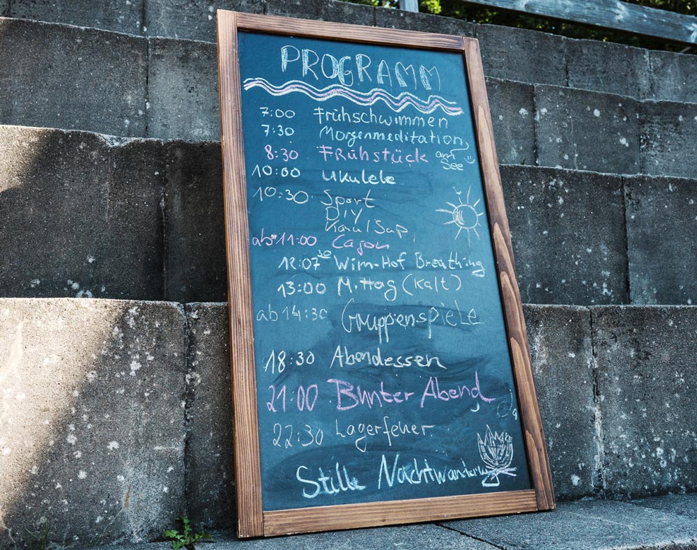 Programmtafel des Sommer-Teamevents mit der Tagesagenda: Frühstück, Sport und Lagerfeuer.